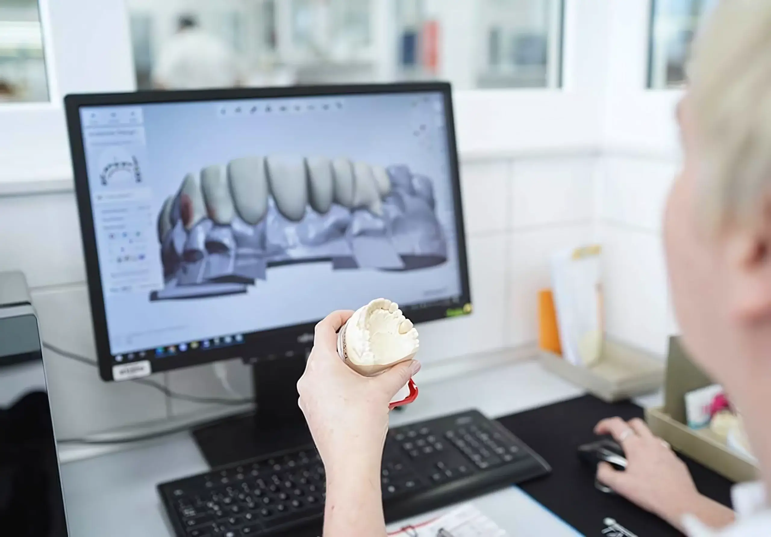 Eine Person vergleicht ein digitales Zahnmodell auf dem Bildschirm mit einem physischen Modell in der Hand bei Voigt dental, was eine der Aufgaben eines Dentallabors widerspiegelt