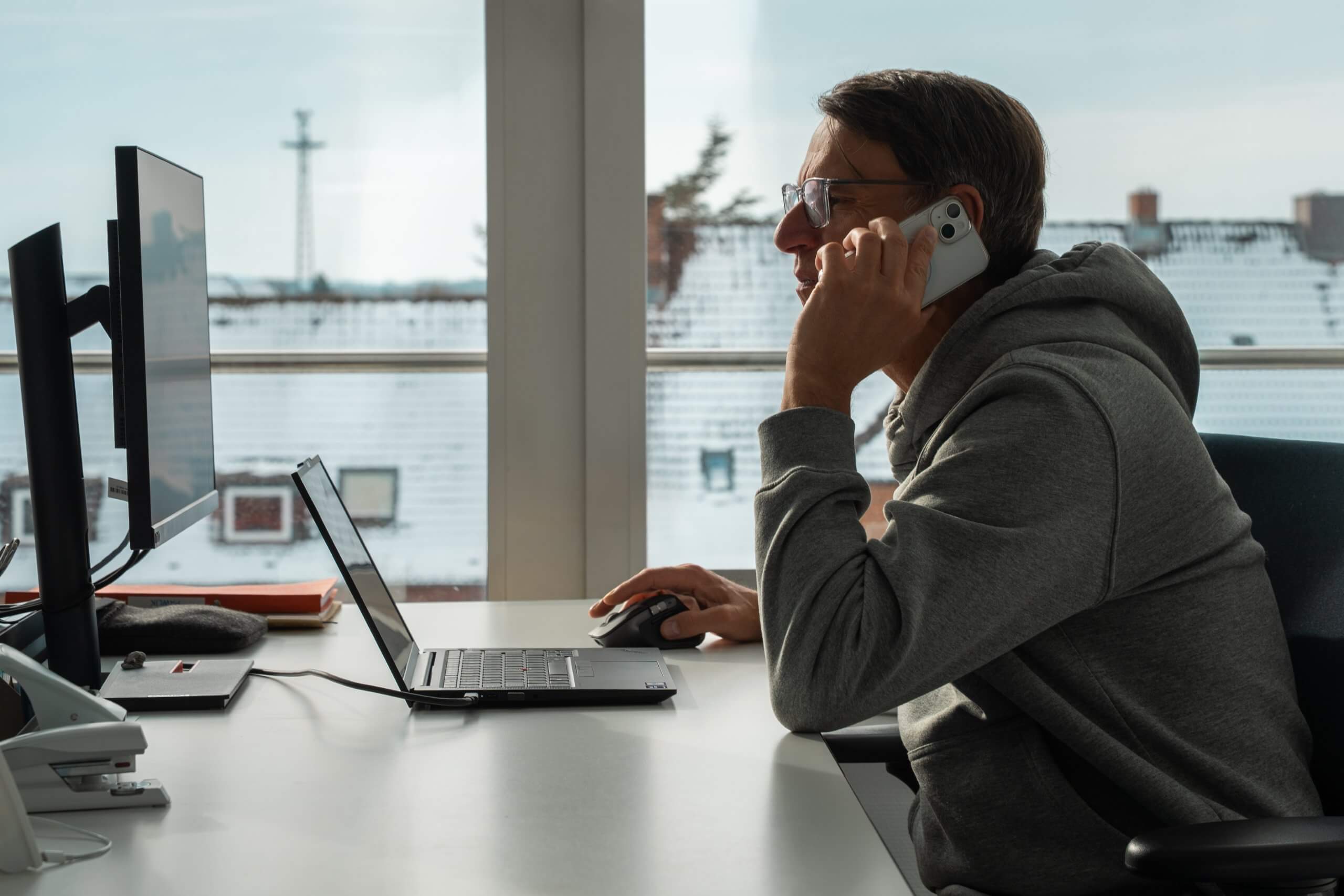 Ein Mann sitzt an einem Schreibtisch, telefoniert und arbeitet am Laptop mit einer Maus, während er aus dem Fenster auf eine verschneite Landschaft blickt.